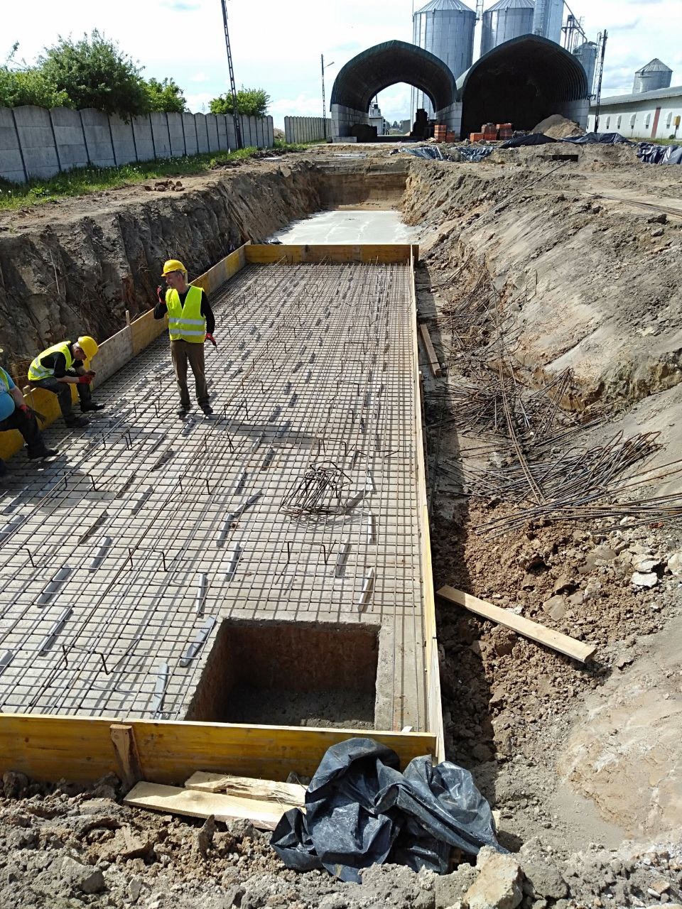 Zbrojenie fundamentu pod budynek przemysłowy, widoczne szalunki i stalowe pręty tworzące siatkę. Pracownicy w kaskach i kamizelkach odblaskowych nadzorują prace.