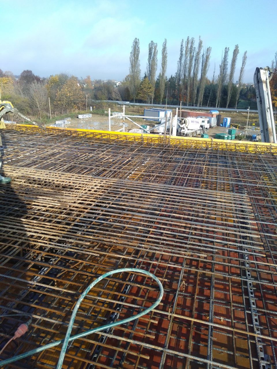 Zbrojenie stropu z widoczną konstrukcją szalunkową, na tle krajobrazu z drzewami i budynkami. Widoczny mikser do betonu i inne maszyny budowlane.