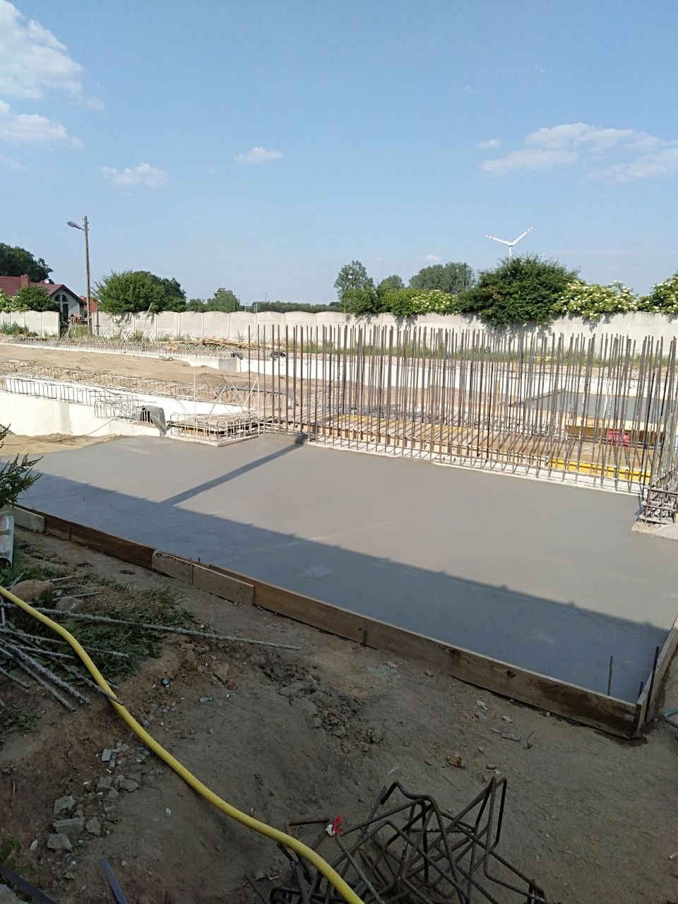 Świeżo wylana, gładka posadzka betonowa z widocznym zbrojeniem pionowym w tle, przygotowana do dalszych prac budowlanych na tle błękitnego nieba.