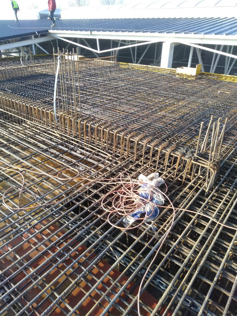 Siatka zbrojeniowa na budowie, widok z góry. Na pierwszym planie zwój drutu wiązałkowego. W tle konstrukcja dachu i pracownicy na nim.