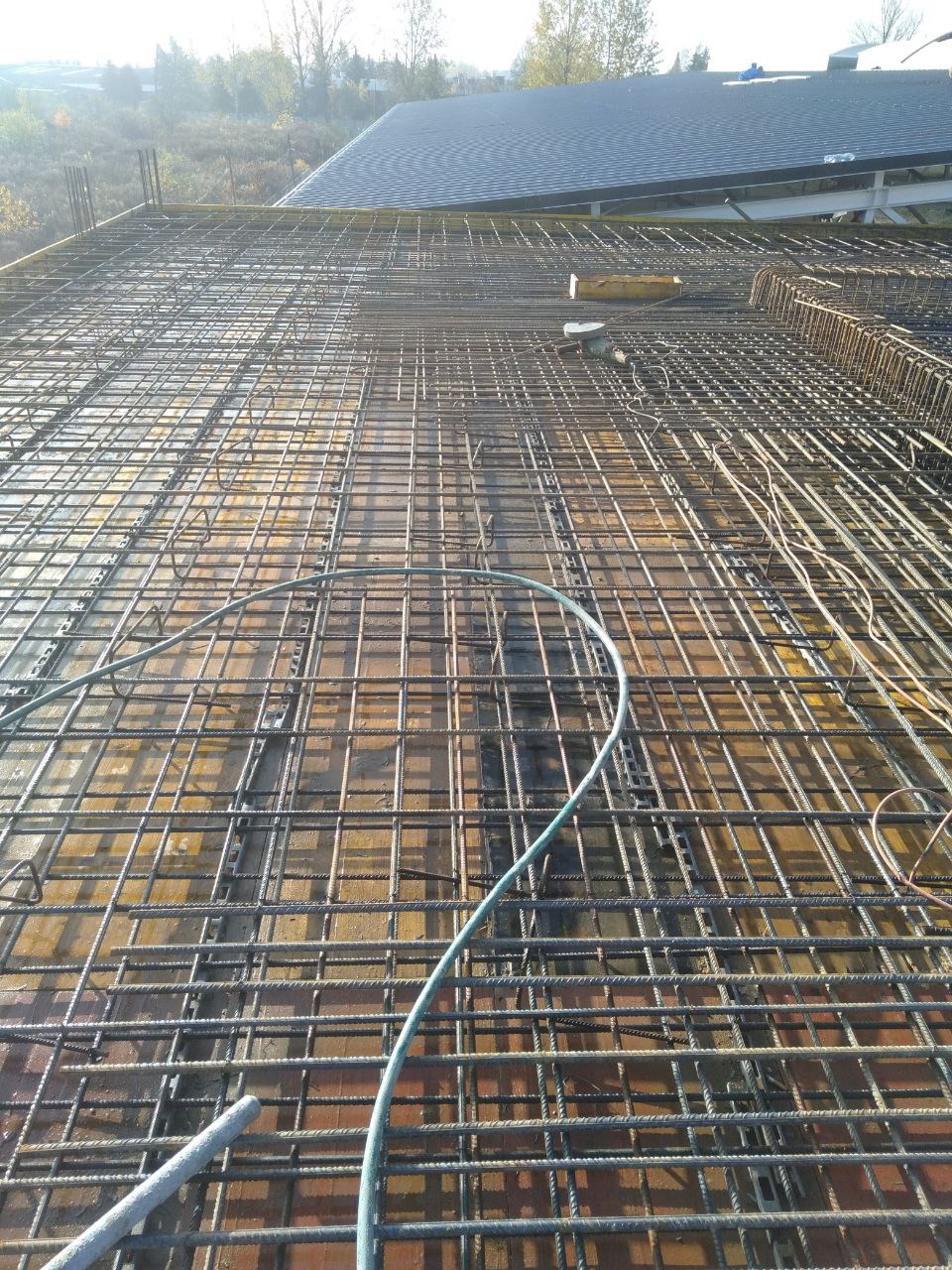 Siatka zbrojeniowa na deskowaniu, widok z góry. Elementy konstrukcyjne budynku w tle, przewody elektryczne na pierwszym planie. Jasne, naturalne światło.