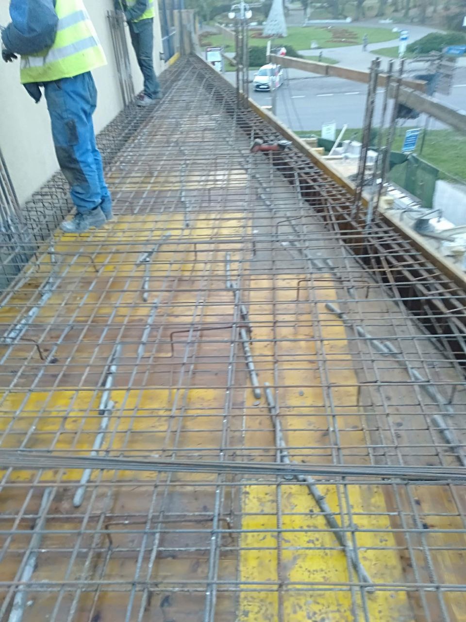 Siatka zbrojeniowa na żółtym szalunku, widok z góry. Dwóch pracowników w oddali. Element konstrukcji budynku w trakcie budowy.