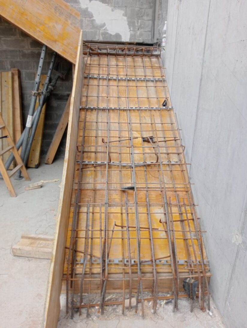 Szalunek zbrojeniowy w narożniku budynku, widoczne deski szalunkowe i siatka zbrojeniowa, przygotowane do zalania betonem. Elementy rusztowania w tle.
