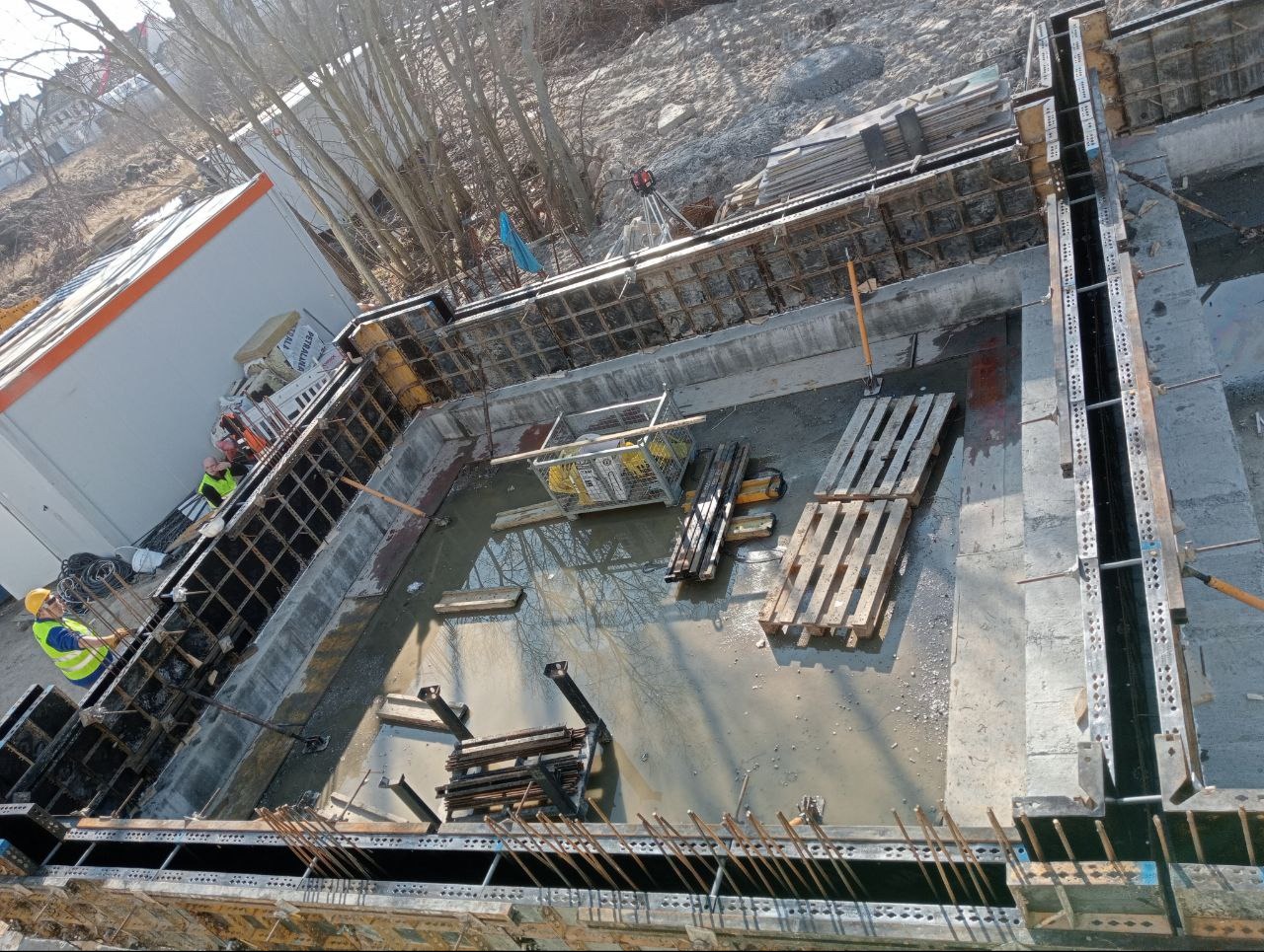 Zalane fundamenty z deskowaniem i zbrojeniem, widok z góry. Na budowie obecni pracownicy w odblaskowych kamizelkach i kaskach. Widoczne palety i narzędzia.