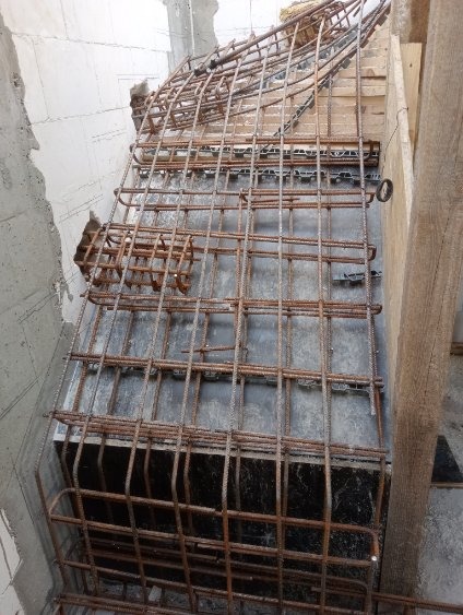 Zbrojenie schodów żelbetowych w szalunku, widok z góry. Elementy zbrojenia połączone drutem. Szalunek z desek i płyt. Surowe ściany w tle.