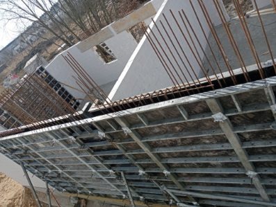 Szare szalunki stropowe z wystającymi prętami zbrojeniowymi, widok z dołu na budowę domu. Elementy konstrukcyjne w surowym stanie.