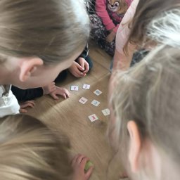 Weronika Ratajczyk - Dzieci siedzą na podłodze i bawią się kartami z literami, tworząc słowa. Widok z góry, skupienie na kartach i dłoniach dzieci. Jasne oświetlenie.