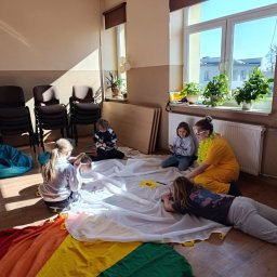 Weronika Ratajczyk - Warsztaty artystyczne dla dzieci w Nowej Suchej. Grupa dzieci i animatorka w żółtej sukience rysują na białym materiale z tęczowym brzegiem. Jasne wnętrze, słoneczny dzień.