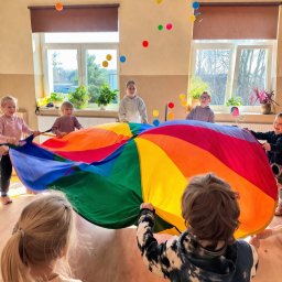 Weronika Ratajczyk - Grupka dzieci bawi się kolorową chustą animacyjną w sali, podrzucając w górę małe piłeczki. W tle okna z roślinami doniczkowymi.