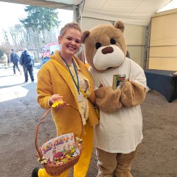 Weronika Ratajczyk - Uśmiechnięta kobieta w żółtym stroju z koszem słodyczy i pluszowy miś z puszką, prawdopodobnie podczas akcji charytatywnej dla dzieci.