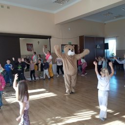 Weronika Ratajczyk - Animator w kostiumie misia tańczy z grupą dzieci w sali z drewnianą podłogą. W tle widoczna flaga Polski i okna z białymi firankami.