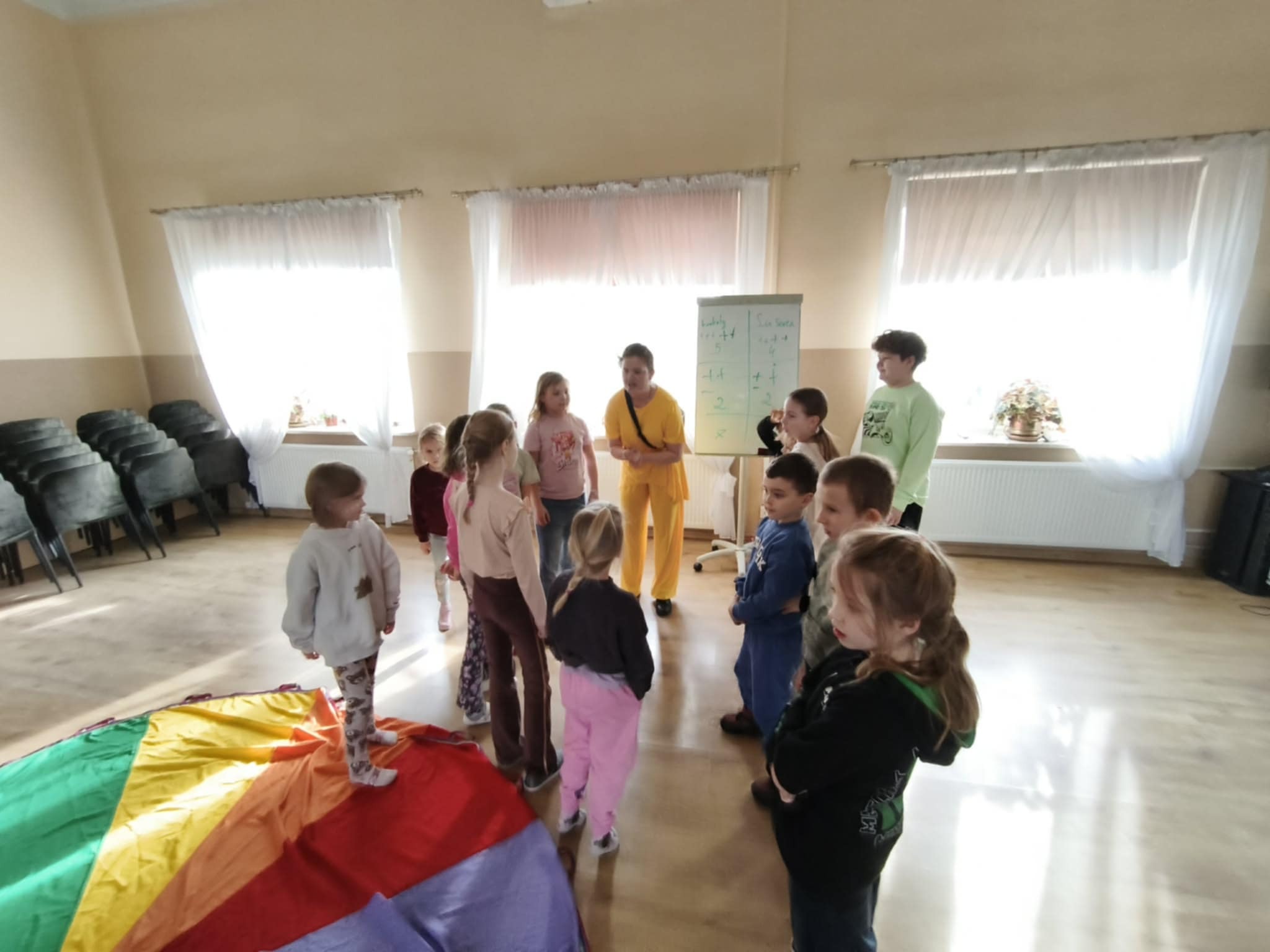 Animator w żółtym stroju prowadzi zabawy z grupą dzieci z kolorową chustą w sali z białymi oknami i krzesłami w tle. Na tablicy zapisane są działania matematyczne.