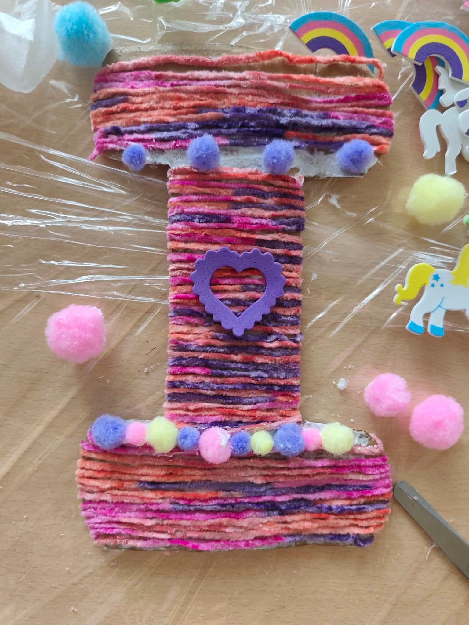 Ręcznie robiona litera 'I' z włóczki w odcieniach różu i fioletu, ozdobiona pomponami i serduszkiem, otoczona elementami dekoracyjnymi na stole.