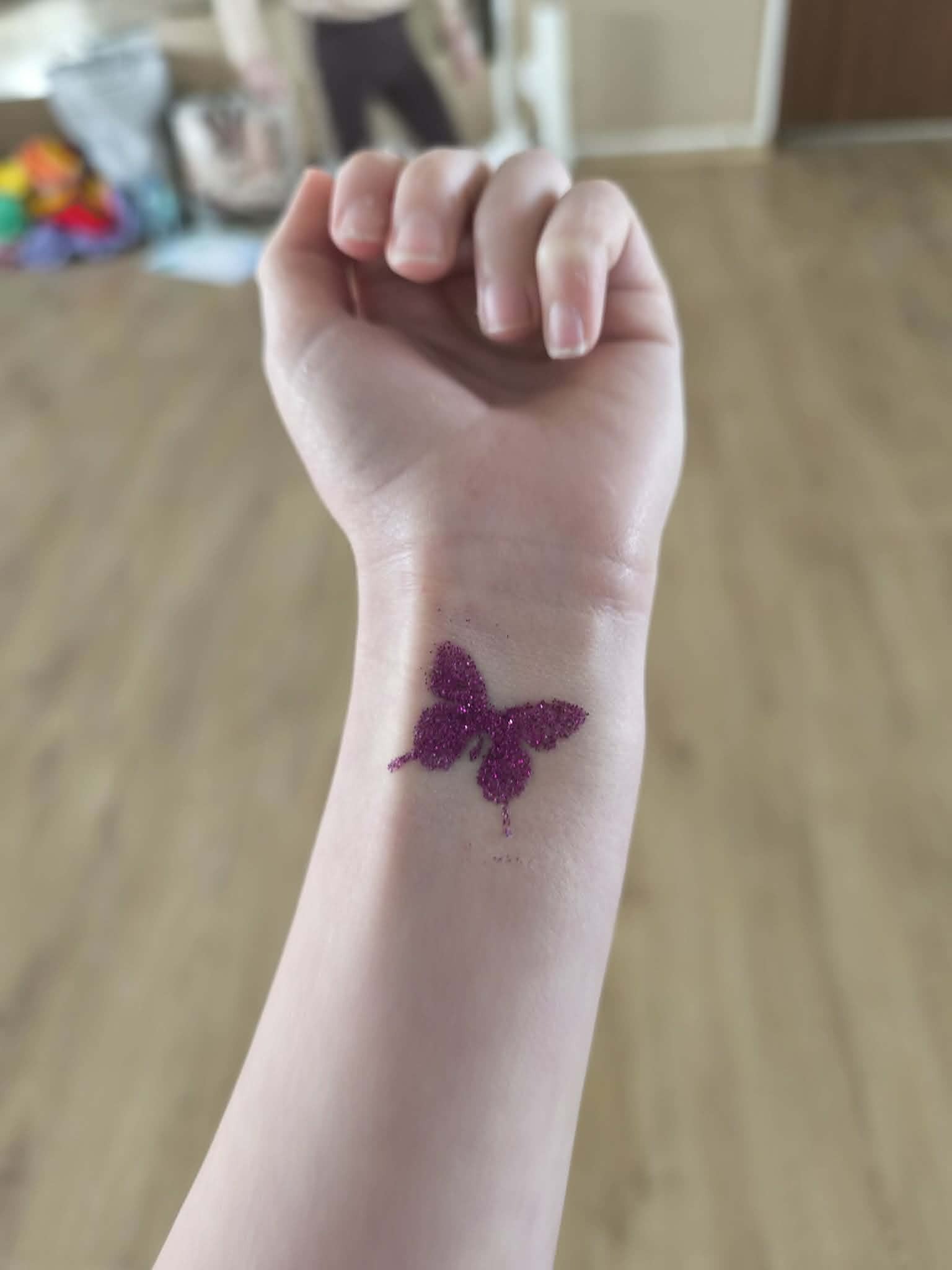 Brokatowy tatuaż w kształcie motyla na nadgarstku osoby, wykonany podczas imprezy dla dzieci. Fioletowy brokat na jasnej skórze, widok z góry.