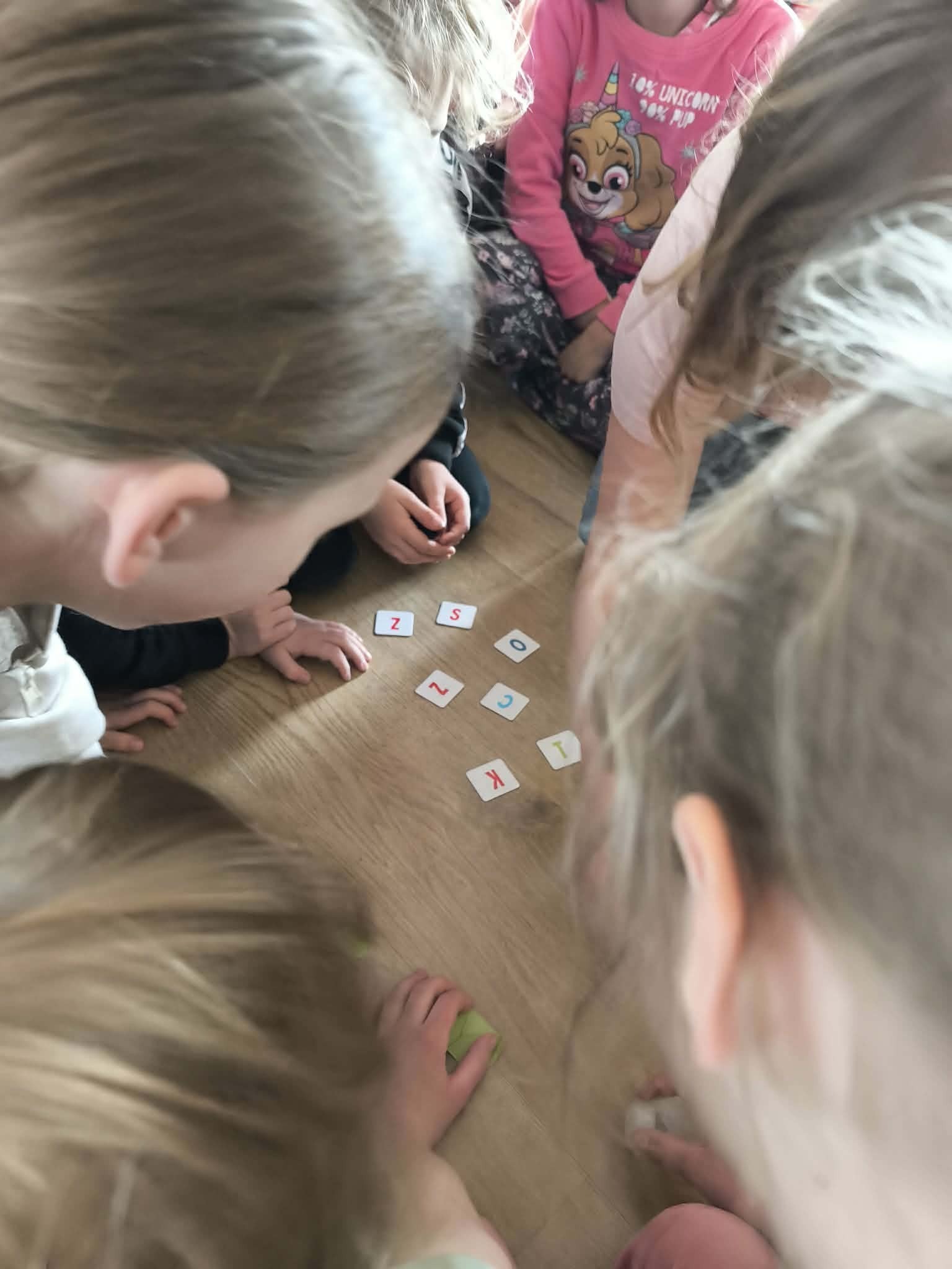 Dzieci siedzą na podłodze i bawią się kartami z literami, tworząc słowa. Widok z góry, skupienie na kartach i dłoniach dzieci. Jasne oświetlenie.
