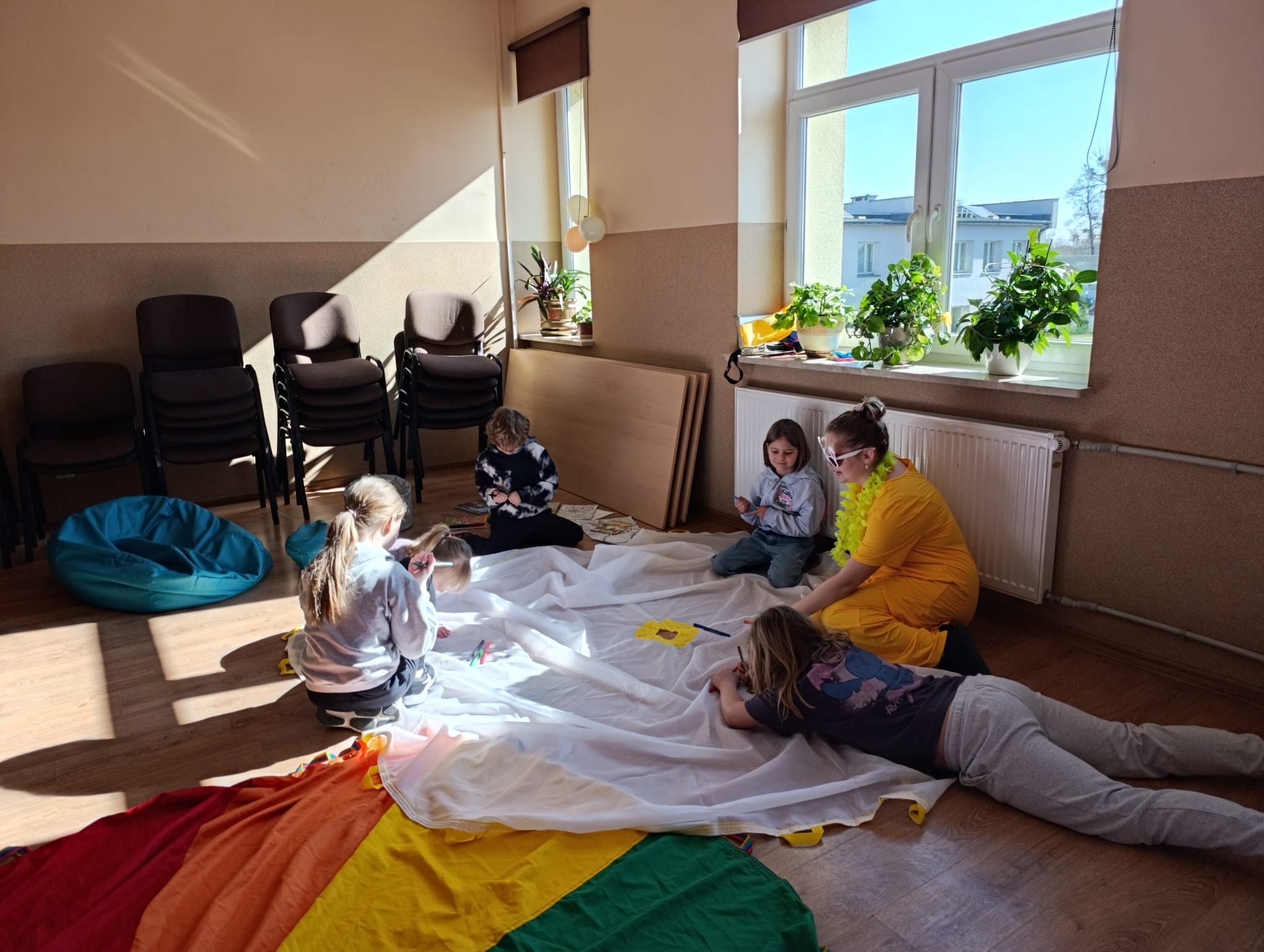 Warsztaty artystyczne dla dzieci w Nowej Suchej. Grupa dzieci i animatorka w żółtej sukience rysują na białym materiale z tęczowym brzegiem. Jasne wnętrze, słoneczny dzień.