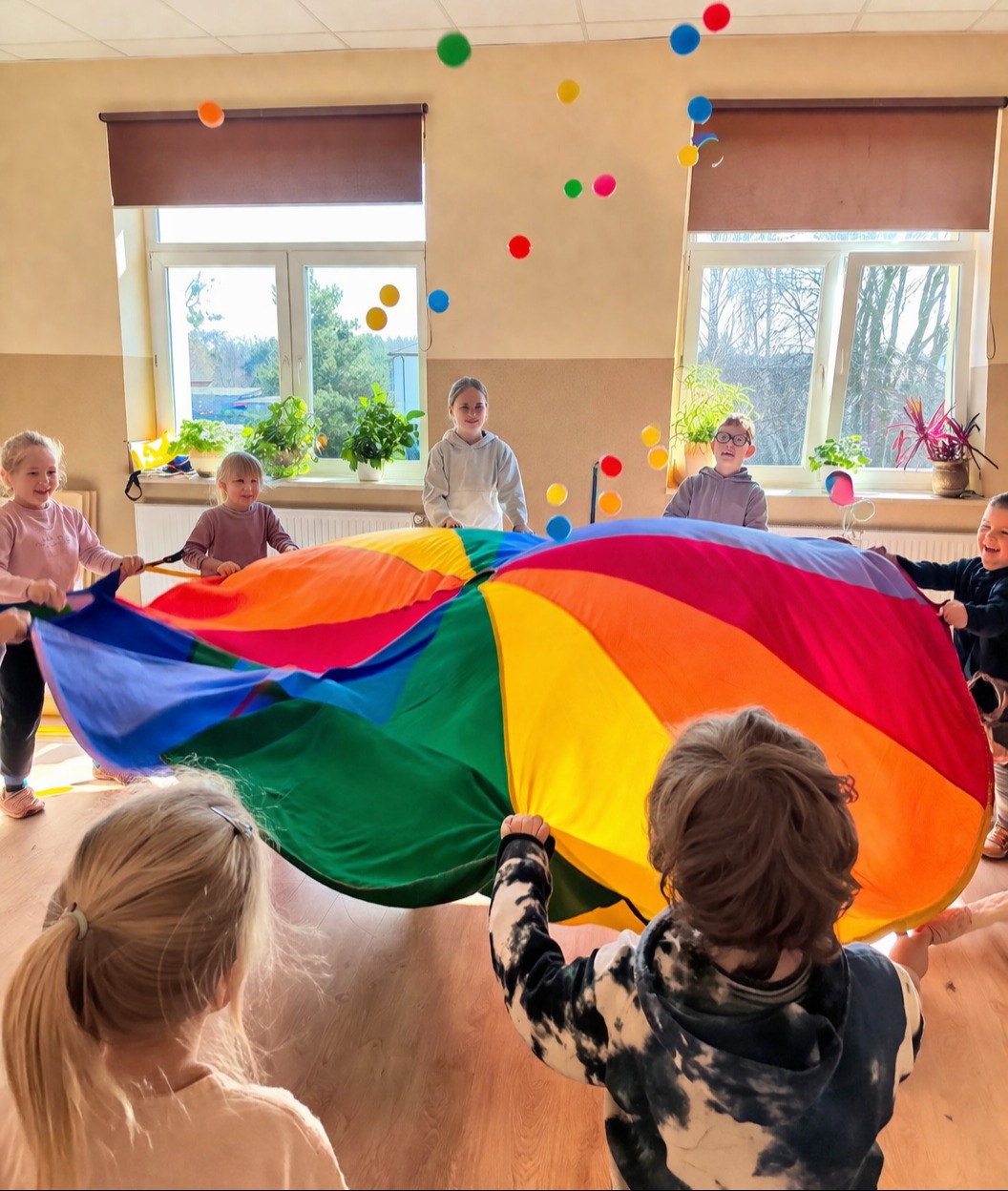 Grupka dzieci bawi się kolorową chustą animacyjną w sali, podrzucając w górę małe piłeczki. W tle okna z roślinami doniczkowymi.