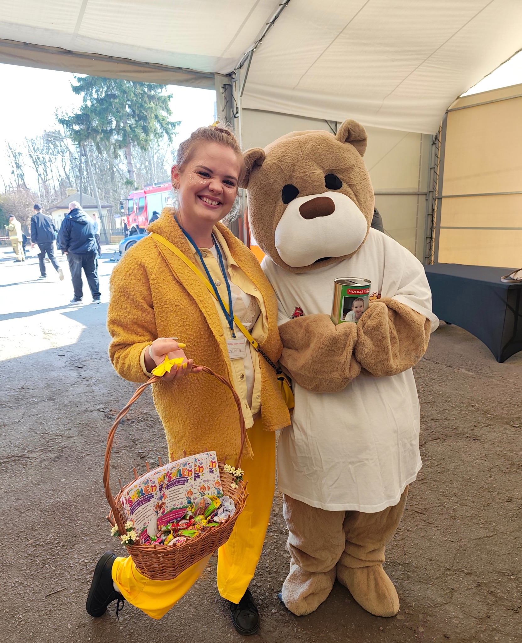 Uśmiechnięta kobieta w żółtym stroju z koszem słodyczy i pluszowy miś z puszką, prawdopodobnie podczas akcji charytatywnej dla dzieci.
