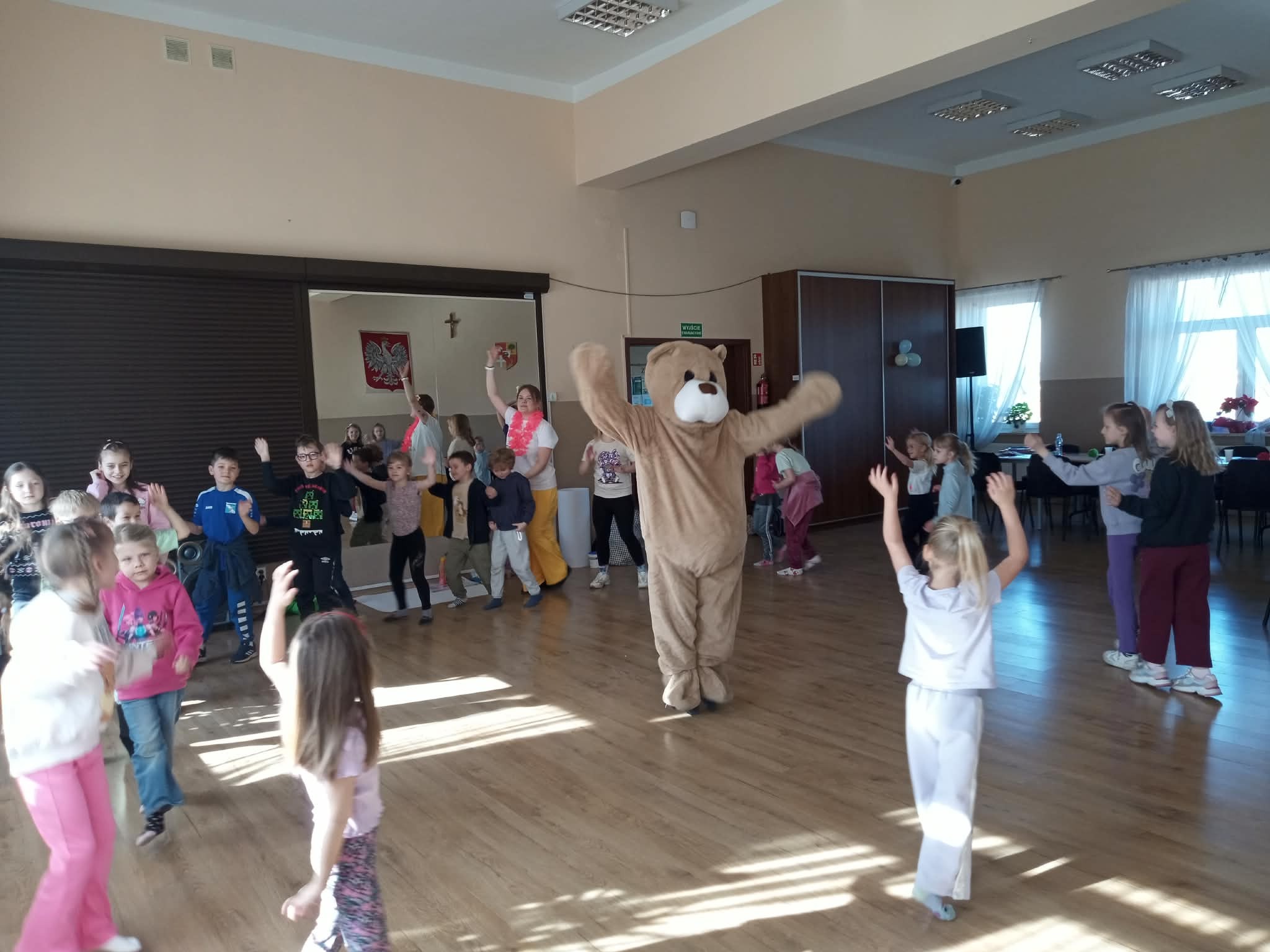 Animator w kostiumie misia tańczy z grupą dzieci w sali z drewnianą podłogą. W tle widoczna flaga Polski i okna z białymi firankami.