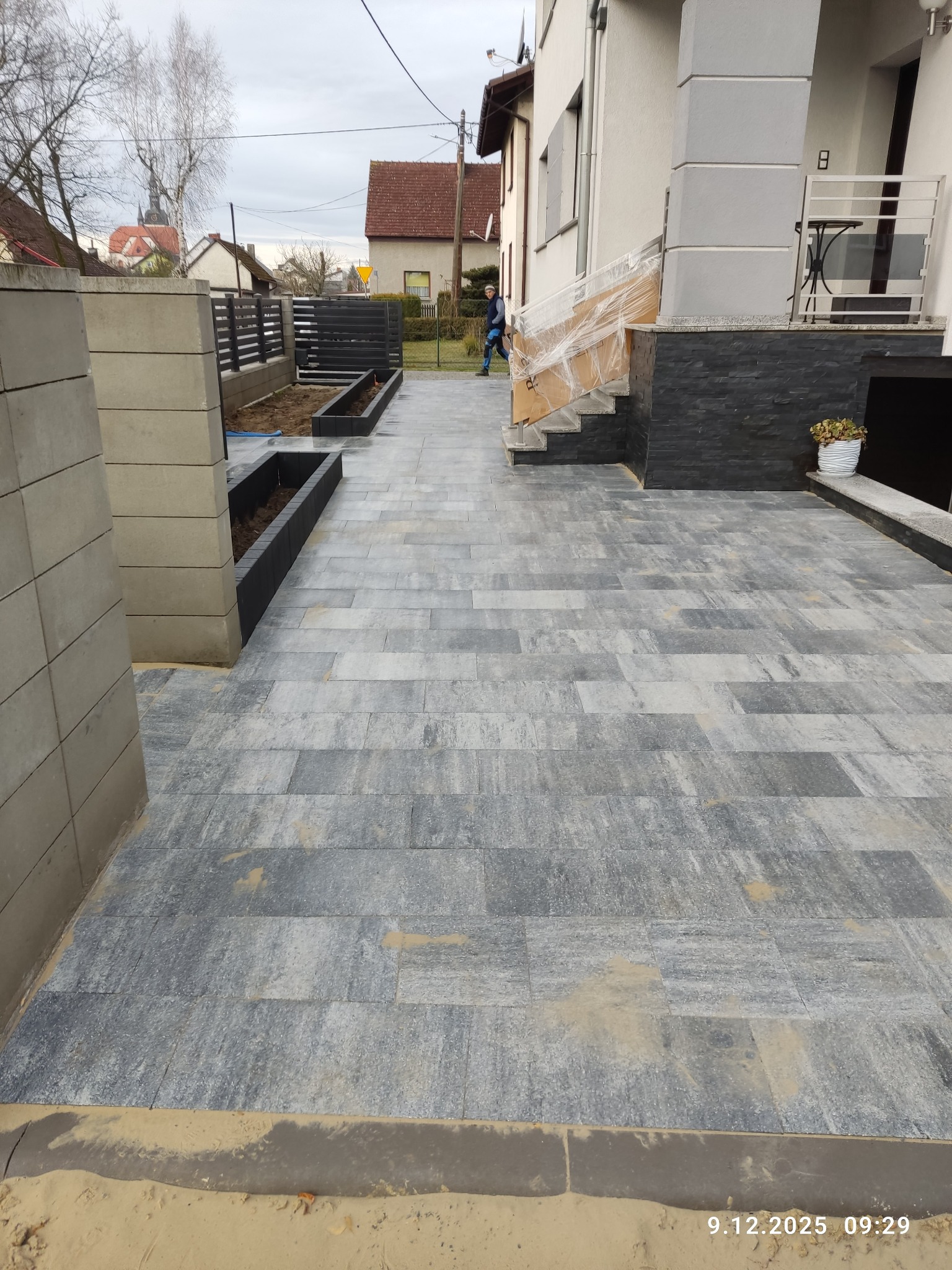 Ułożona szara kostka brukowa na podjeździe, z widocznymi elementami małej architektury: rabaty, ogrodzenie panelowe, schody do domu. W tle zabudowa jednorodzinna.
