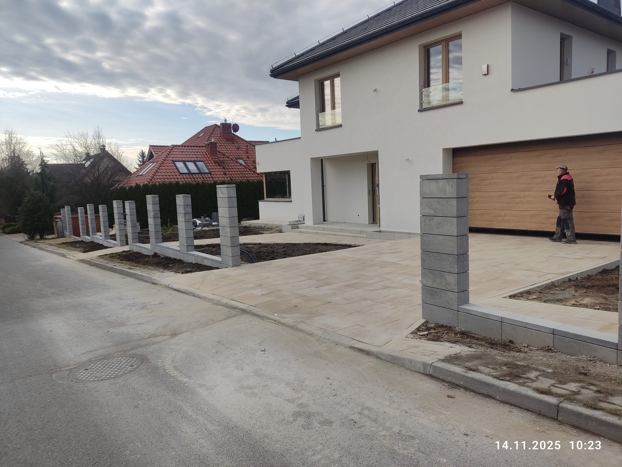 Nowoczesny podjazd z jasnej kostki brukowej prowadzący do domu z drewnianą bramą garażową. W tle widoczne słupki ogrodzeniowe i fragment dachu sąsiedniego budynku.