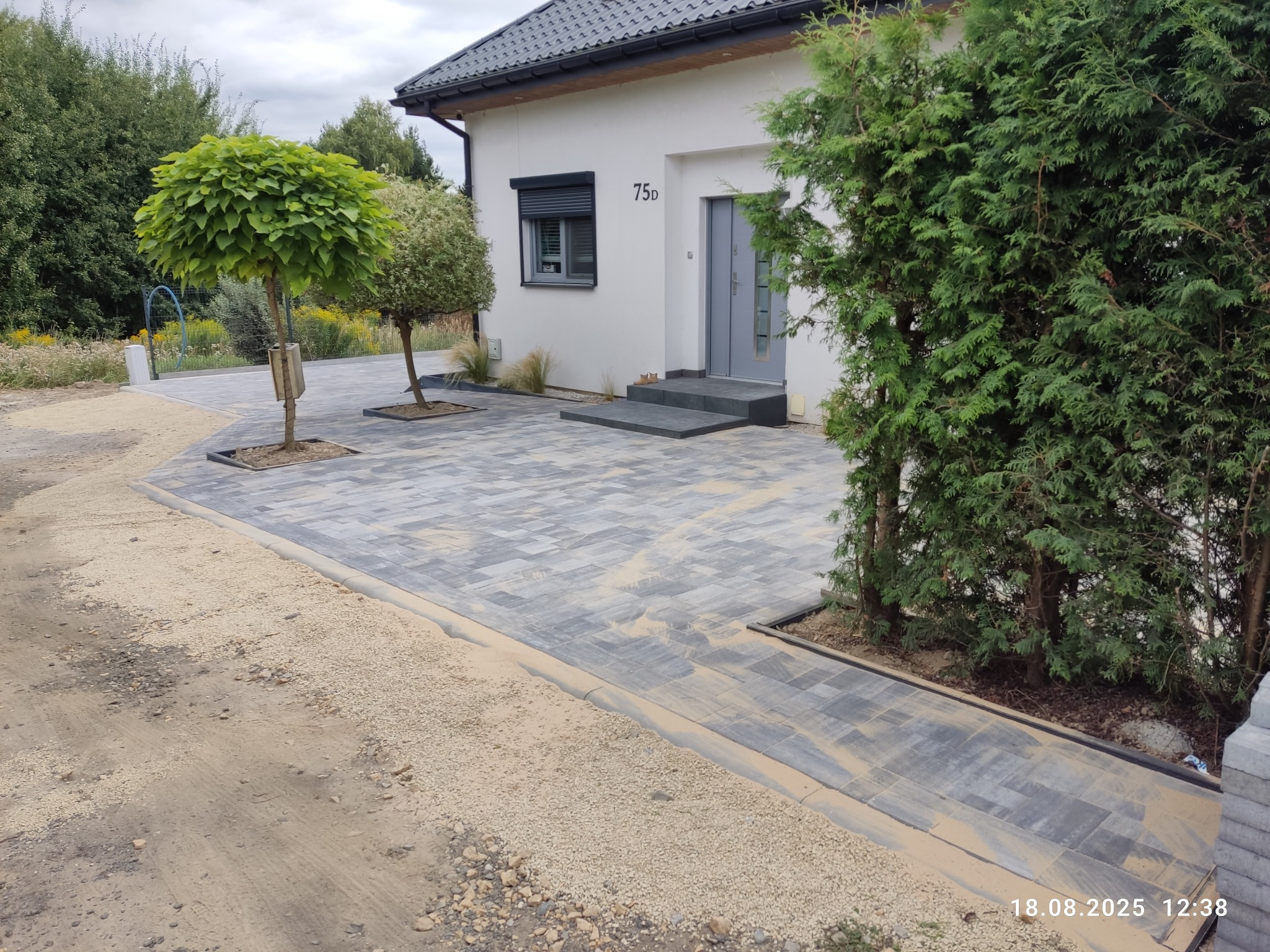 Ułożona kostka brukowa w szaro-beżowej tonacji przed nowoczesnym domem z numerem 75d. Widoczne drzewa i krzewy. Część kostki zasypana piaskiem.