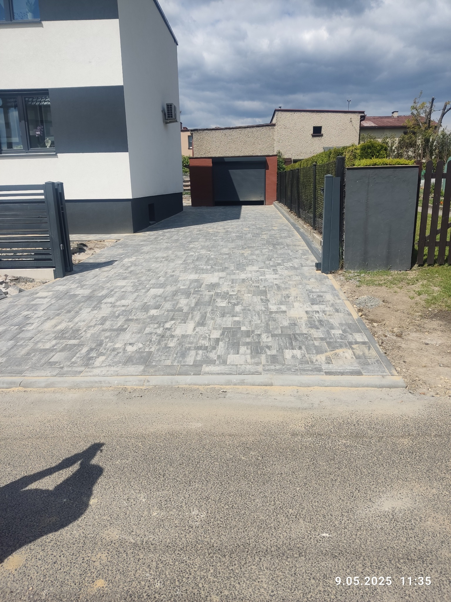 Ułożona szara kostka brukowa prowadząca do garażu, obok nowoczesny dom z szarym ogrodzeniem panelowym i drewnianym. Widoczny cień osoby.