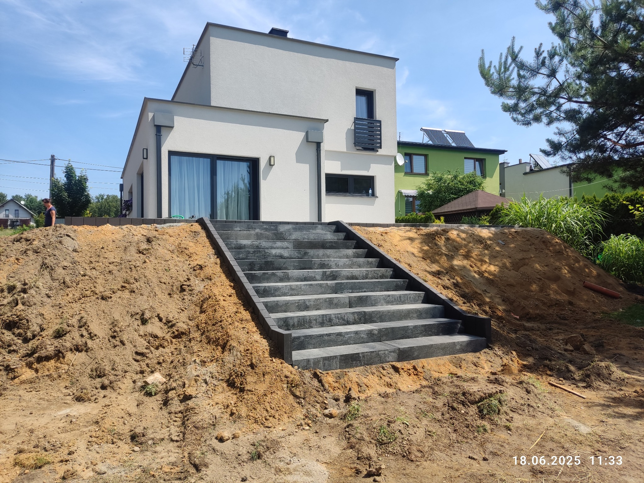Nowoczesne schody z kostki brukowej prowadzące do domu, otoczone ziemią. Widoczny fragment budynku i zieleń w tle. Słoneczny dzień, wyraźne detale.