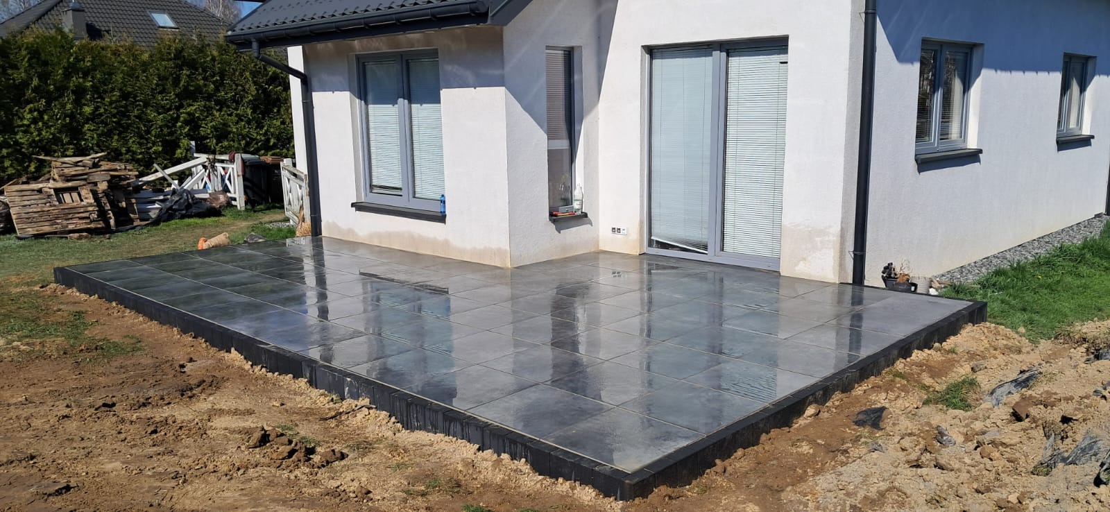 Nowoczesny taras z szarych płyt, obrzeża z czarnej kostki. Widok na biały budynek z szarymi oknami i drzwi balkonowe. W tle zielona roślinność i stos drewna.