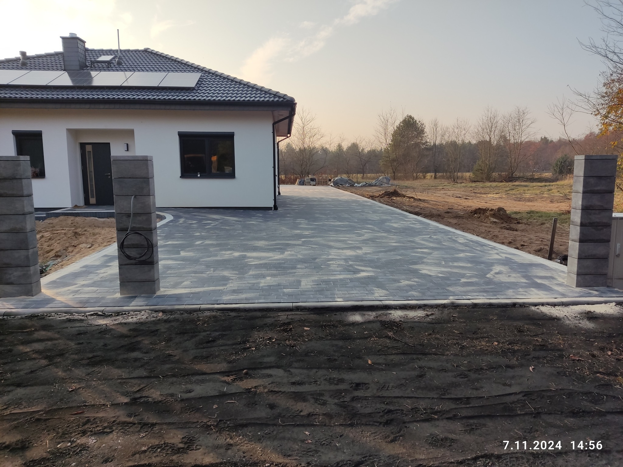 Nowoczesny podjazd z szarej kostki brukowej prowadzący do domu z panelami słonecznymi, wykończony słupkami ogrodzeniowymi z betonu architektonicznego.