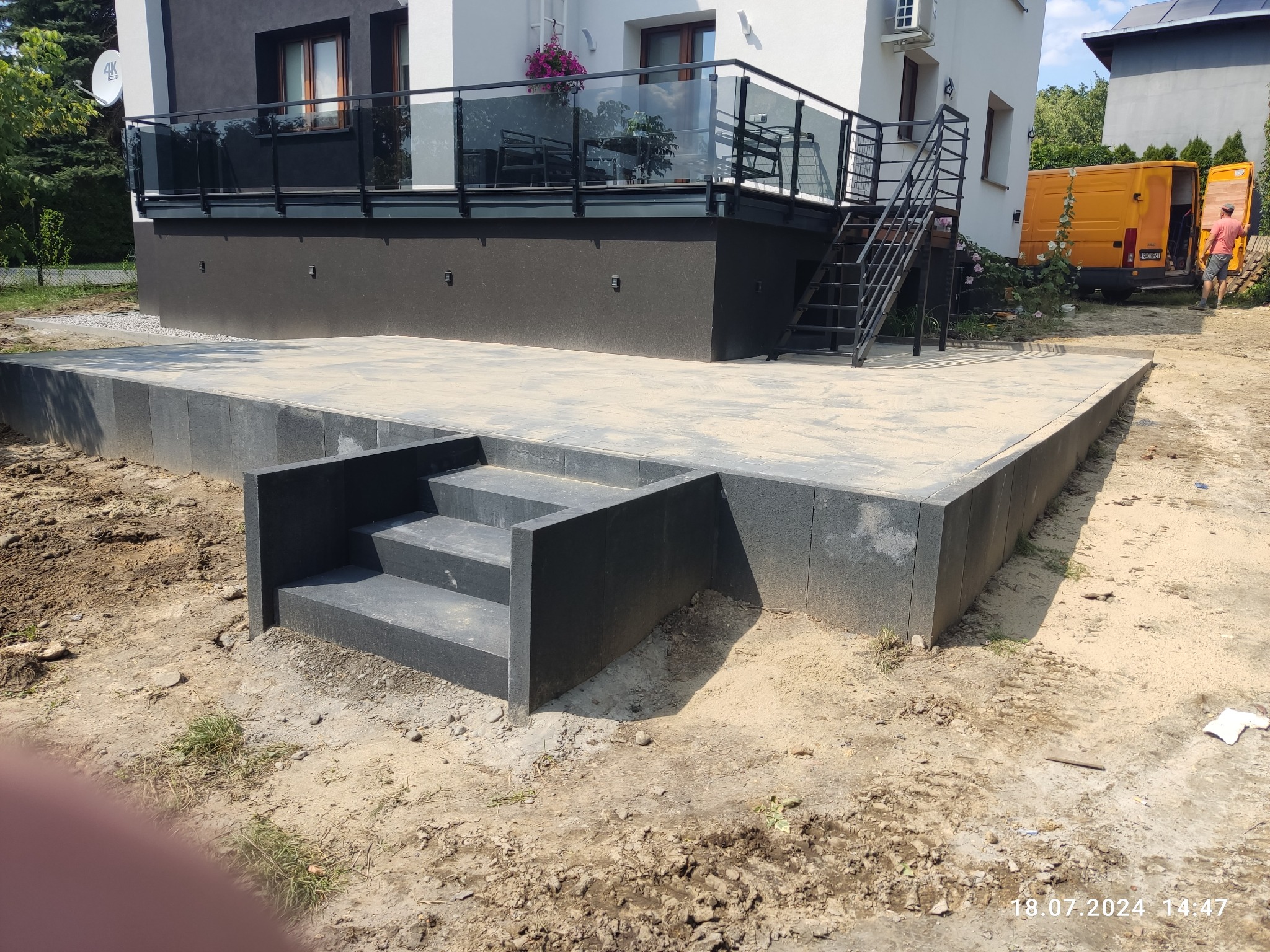 Nowoczesny taras z kostki w odcieniach szarości, z niskimi schodami wejściowymi, przylegający do budynku z balkonem i metalowymi schodami.