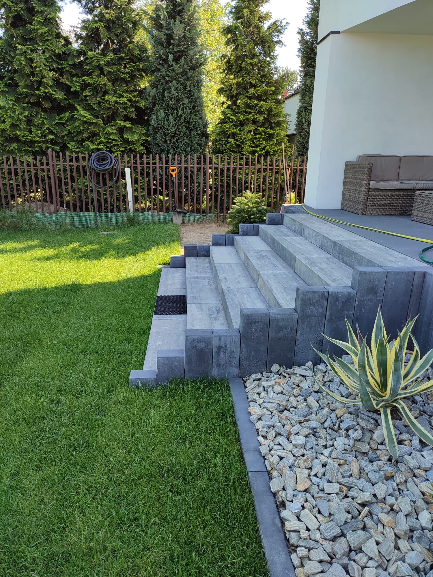 Schody z kostki brukowej prowadzące na taras, obok rabata z kamieni i roślin, w tle zielony trawnik i ogrodzenie z siatki.