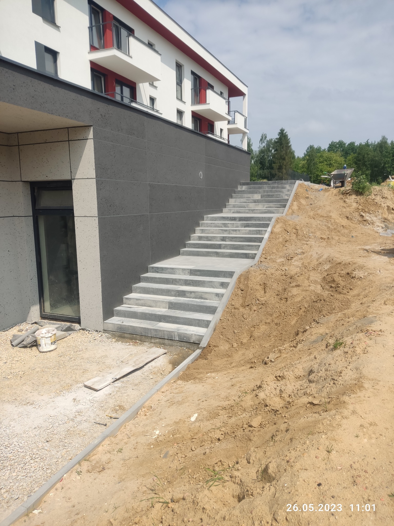 Schody z kostki brukowej prowadzące do budynku, teren wokół w trakcie prac ziemnych. Widoczny fragment budynku z balkonami i okno piwniczne.