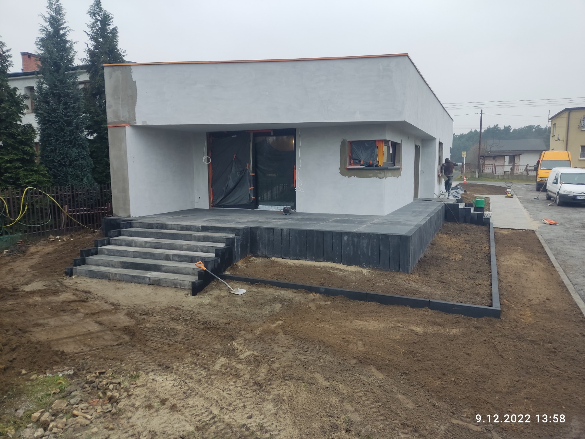 Nowoczesny dom z tarasem wyłożonym ciemną kostką. Schody prowadzą na taras, a obok widoczny fragment ogrodu z ziemią i obrzeżami. Prace wykończeniowe w toku.