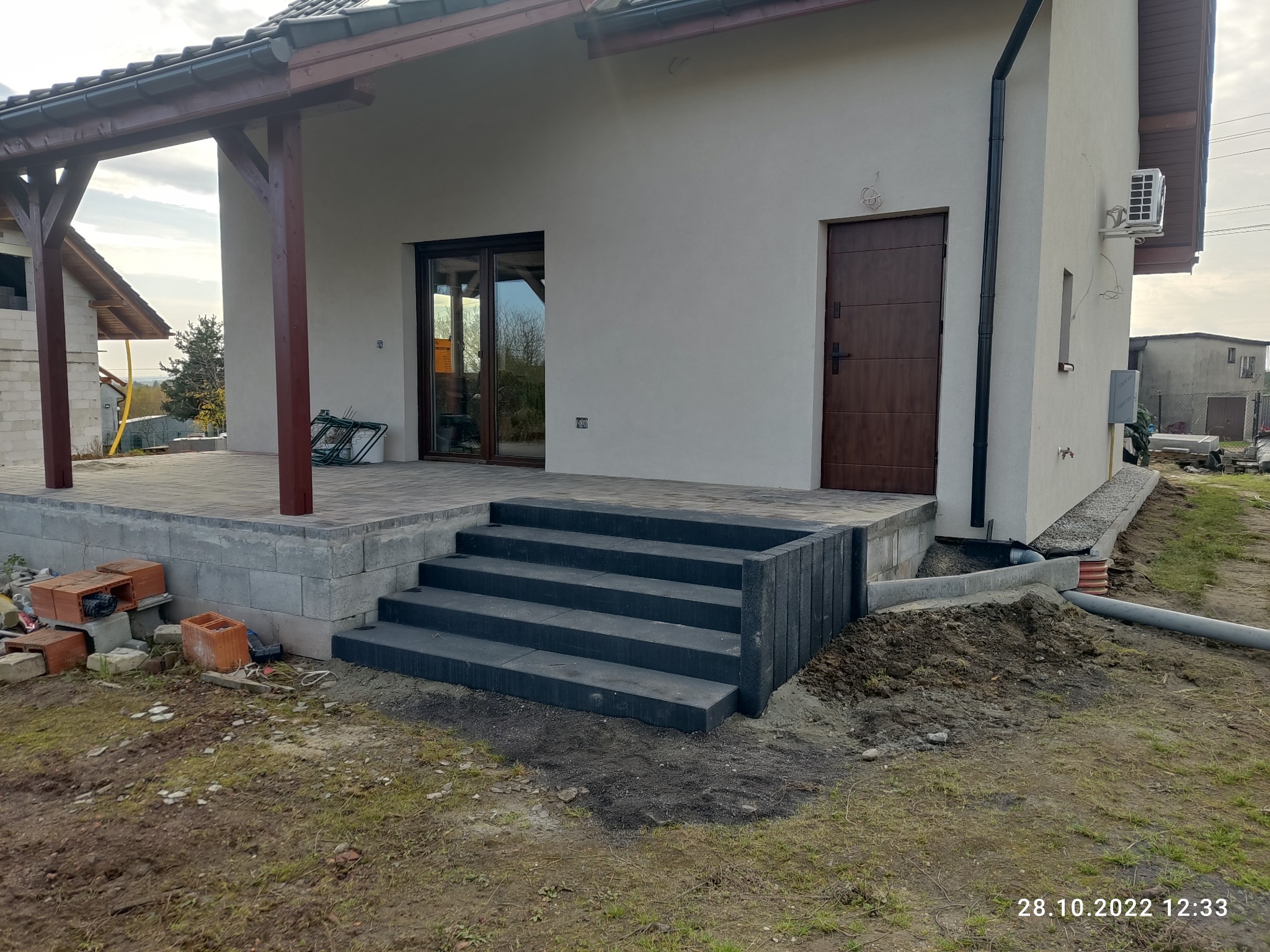 Wykończone schody wejściowe do domu z ciemnej kostki brukowej, z widocznym tarasem z jasnej kostki. Częściowo zagospodarowany teren wokół.