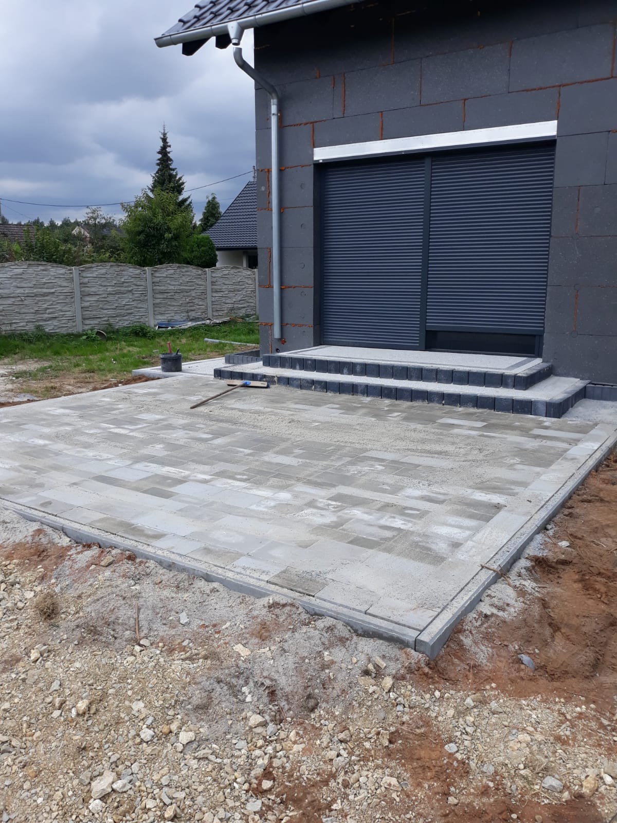 Jasnoszara kostka brukowa ułożona na posesji, widoczne schody z kostki, obrzeża i fragment budynku z szarą elewacją oraz roletami zewnętrznymi. W tle ogrodzenie.