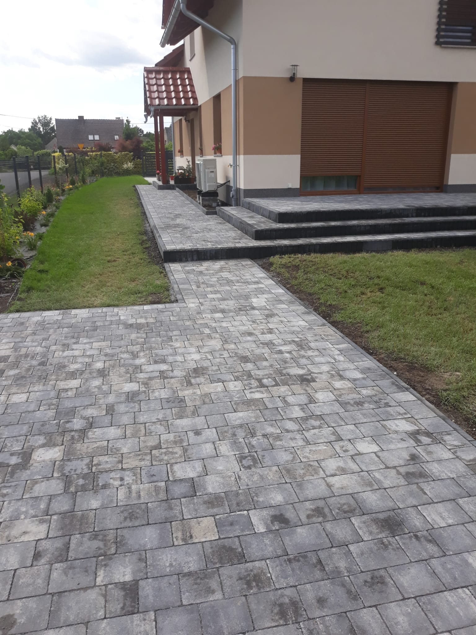 Szara kostka brukowa prowadzi do domu z zielonym trawnikiem i rabatami kwiatowymi. Widoczne schody z kostki i fragment ogrodzenia.