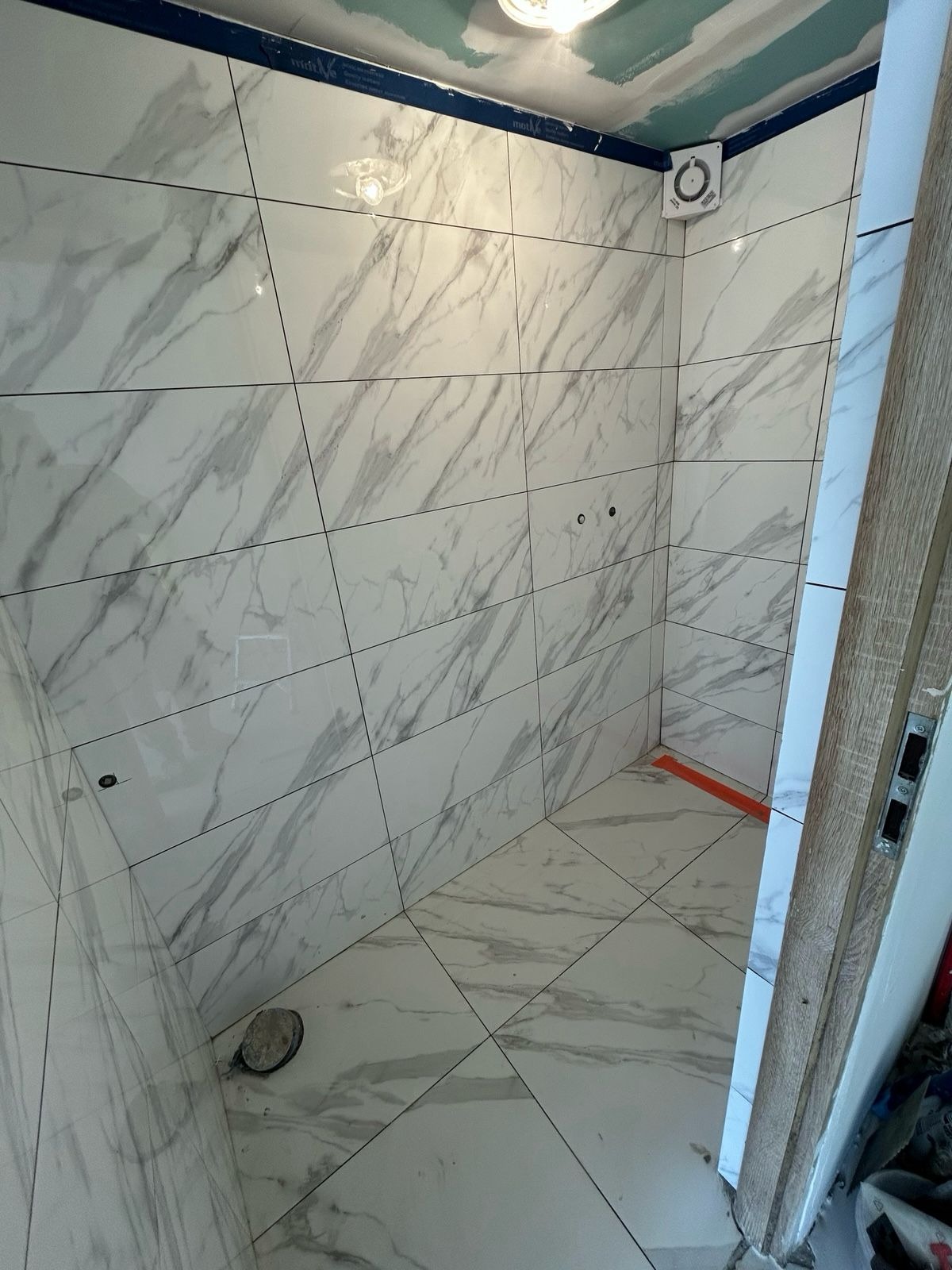 Wykończona łazienka z białymi płytkami imitującymi marmur na ścianach i podłodze. Widoczny fragment drzwi i wentylator. Otwory na baterię.