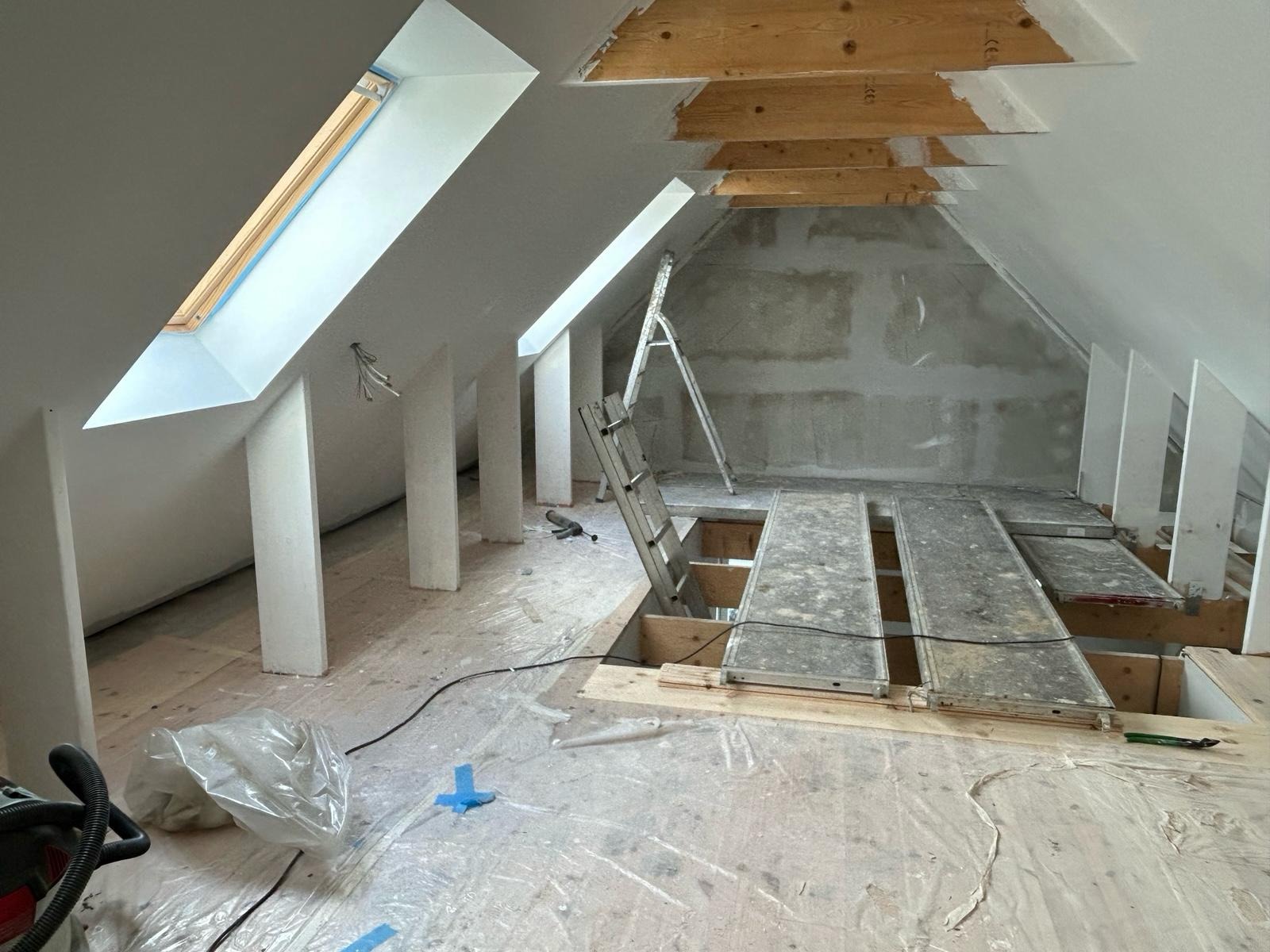 Adaptacja poddasza: widok na ocieplone skosy z oknami dachowymi, rusztowanie i drabina. Prace wykończeniowe w toku, podłoga zabezpieczona folią.