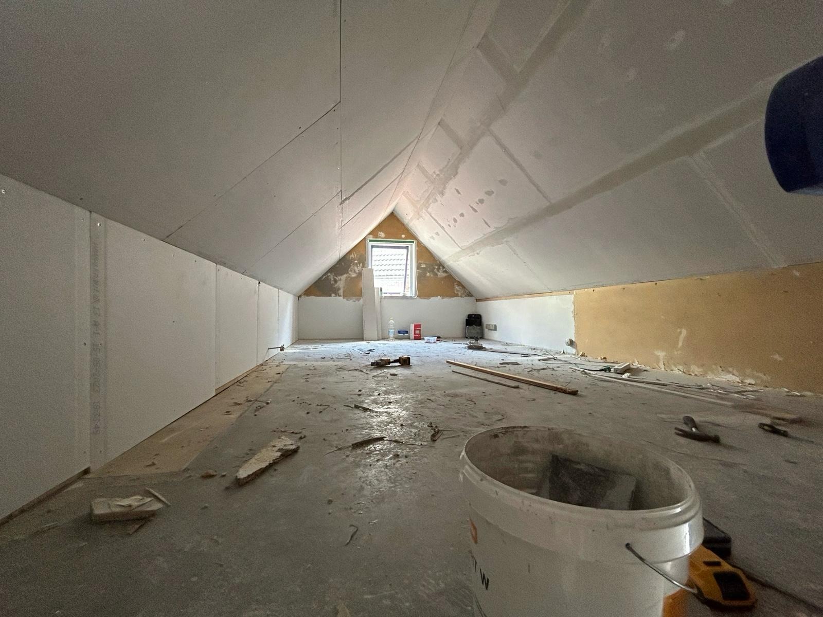 Poddasze w trakcie prac wykończeniowych: ściany obłożone płytami gipsowymi, widoczne narzędzia i wiadro z farbą. Okno w szczycie dachu. Surowy stan, przygotowanie do dalszych prac.