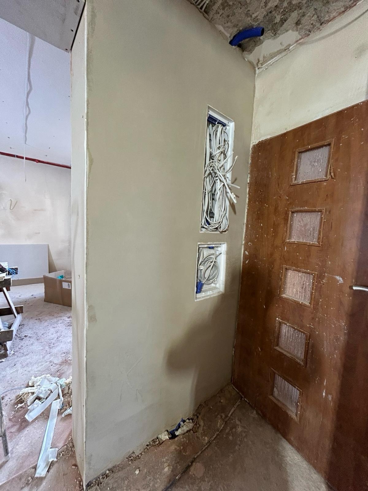 Ściana po szpachlowaniu z otworami na kable elektryczne, widoczna stara, drewniana futryna drzwi. Prace wykończeniowe w trakcie realizacji.