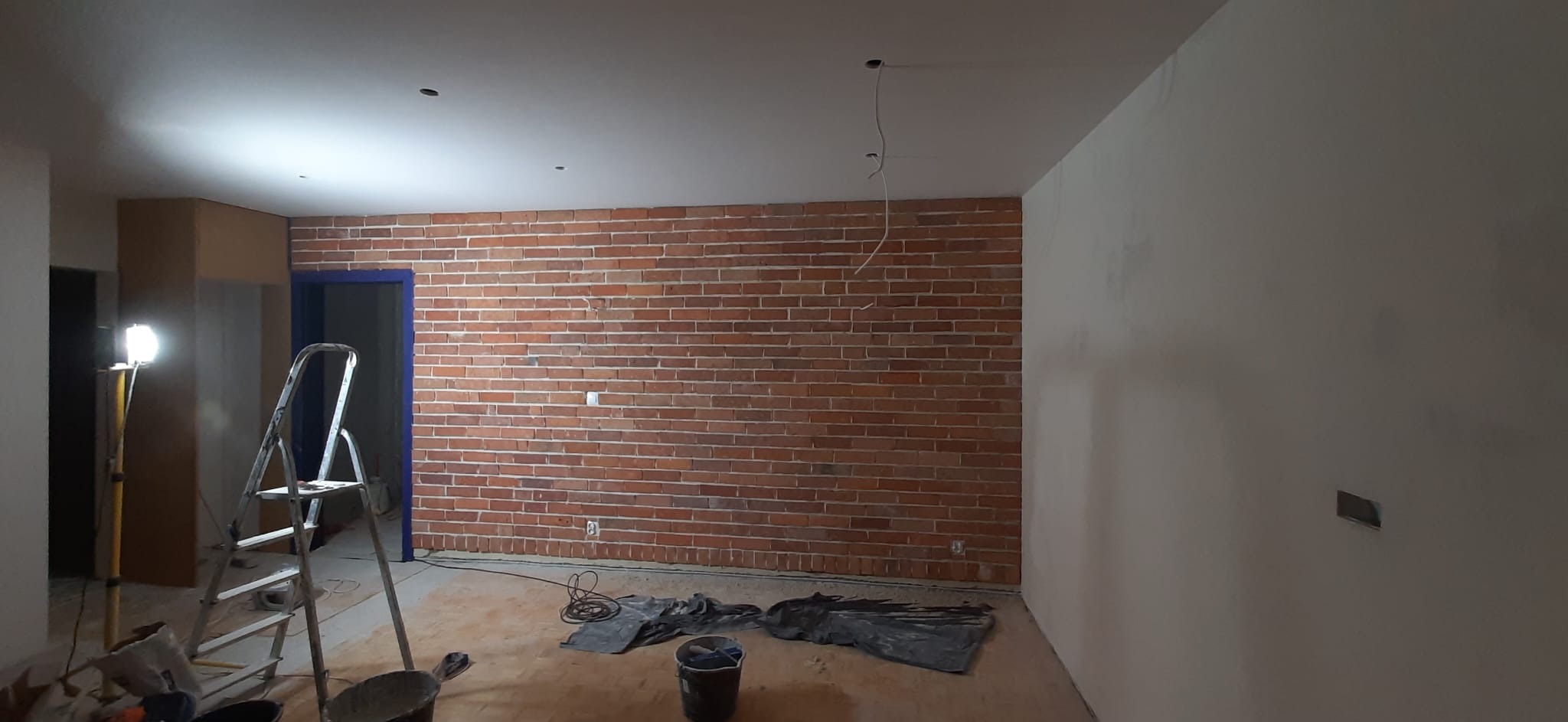 Pokój w trakcie prac wykończeniowych: ściana z cegły, częściowo wyszpachlowana ściana, drabina, kable zwisające z sufitu. Widoczne narzędzia i materiały budowlane.