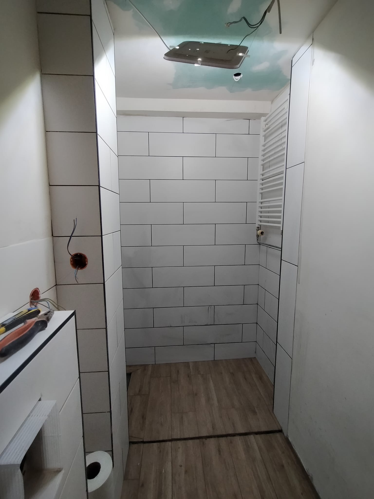 Wykończona łazienka z białymi, prostokątnymi płytkami ułożonymi poziomo, drewnopodobną podłogą i grzejnikiem drabinkowym. Widoczne narzędzia i puszka elektryczna.