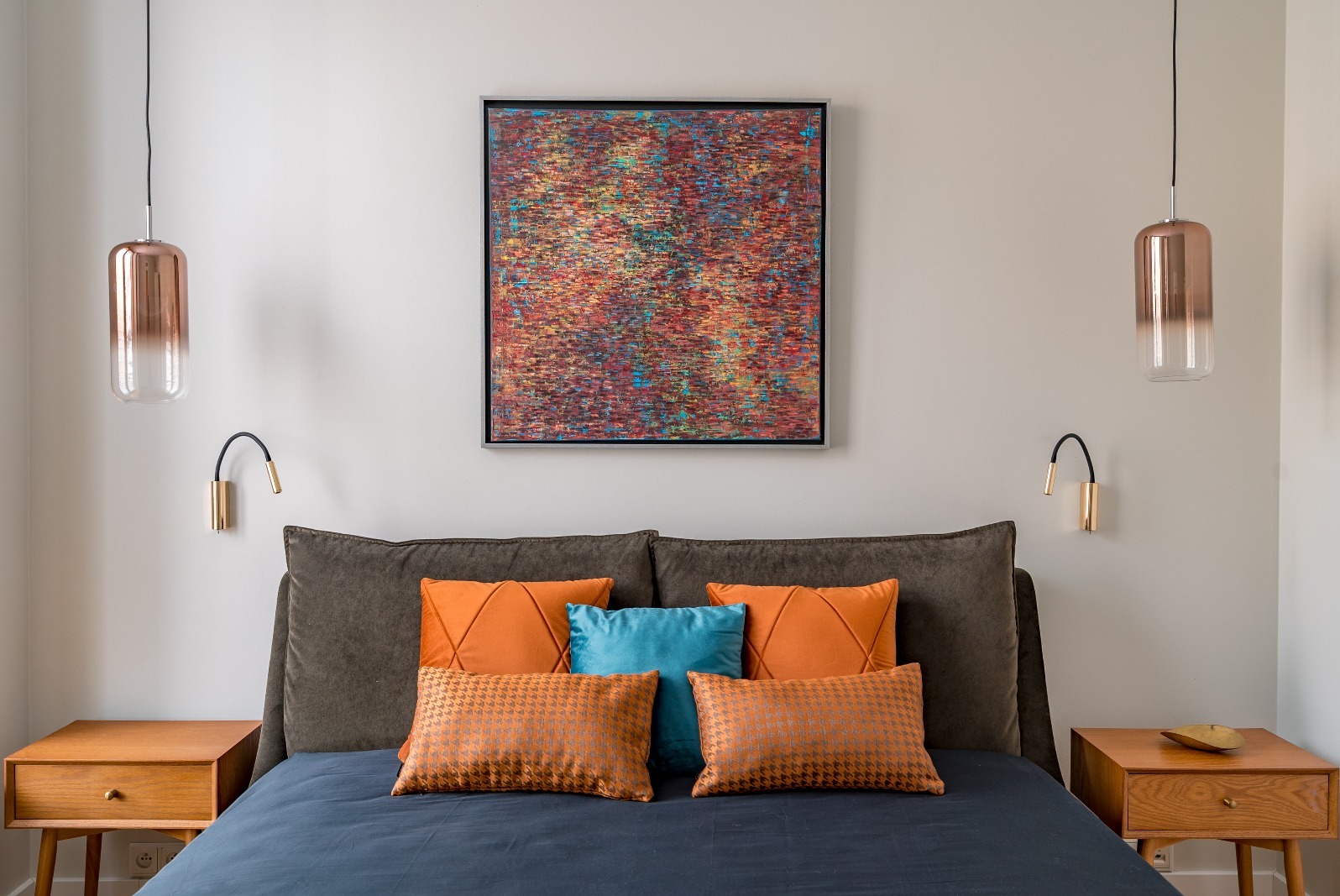 Sypialnia z łóżkiem w odcieniach szarości i granatu, ożywiona pomarańczowymi poduszkami. Nad łóżkiem abstrakcyjny obraz i lampki nocne w stylu art deco.