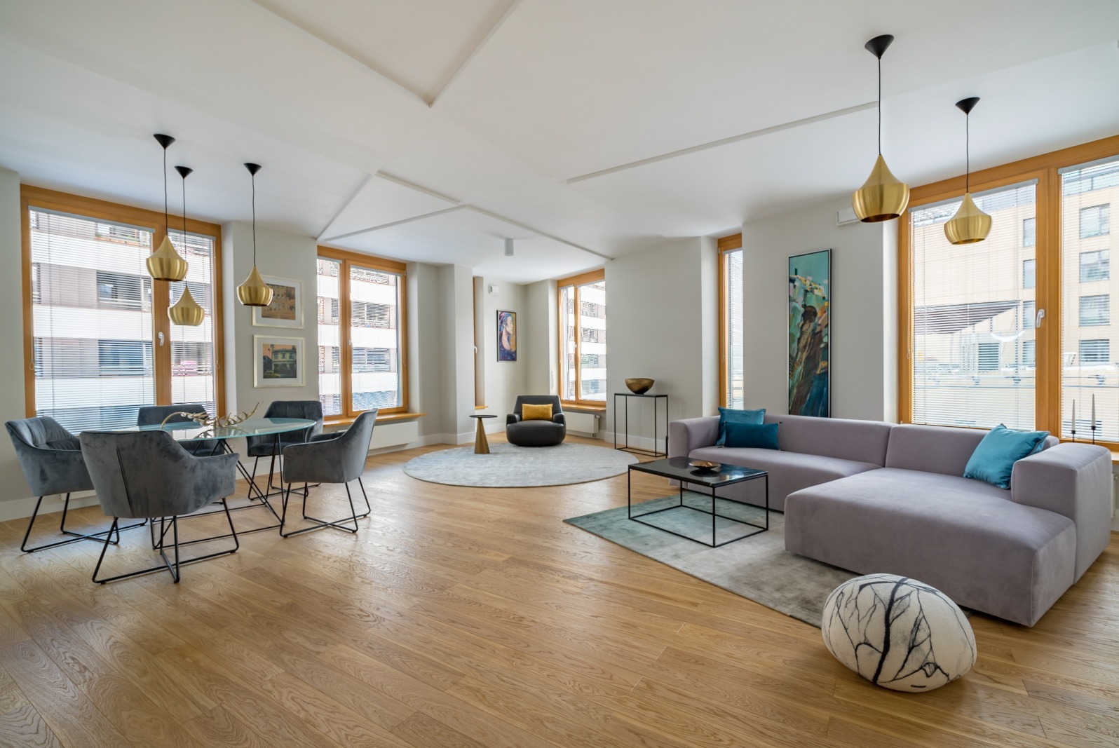 Jasne, przestronne wnętrze z drewnianą podłogą, szarą sofą z turkusowymi poduszkami, okrągłym stołem z krzesłami i złotymi lampami wiszącymi. Nowoczesny design.