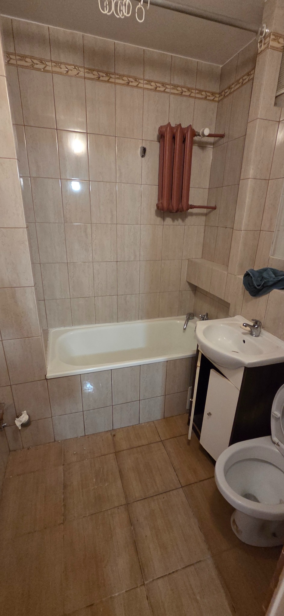 Ujęcie łazienki z beżowymi płytkami, wanną, umywalką z szafką i toaletą. Na ścianie grzejnik żeberkowy. Widoczne ślady użytkowania i brak nowoczesnego designu.