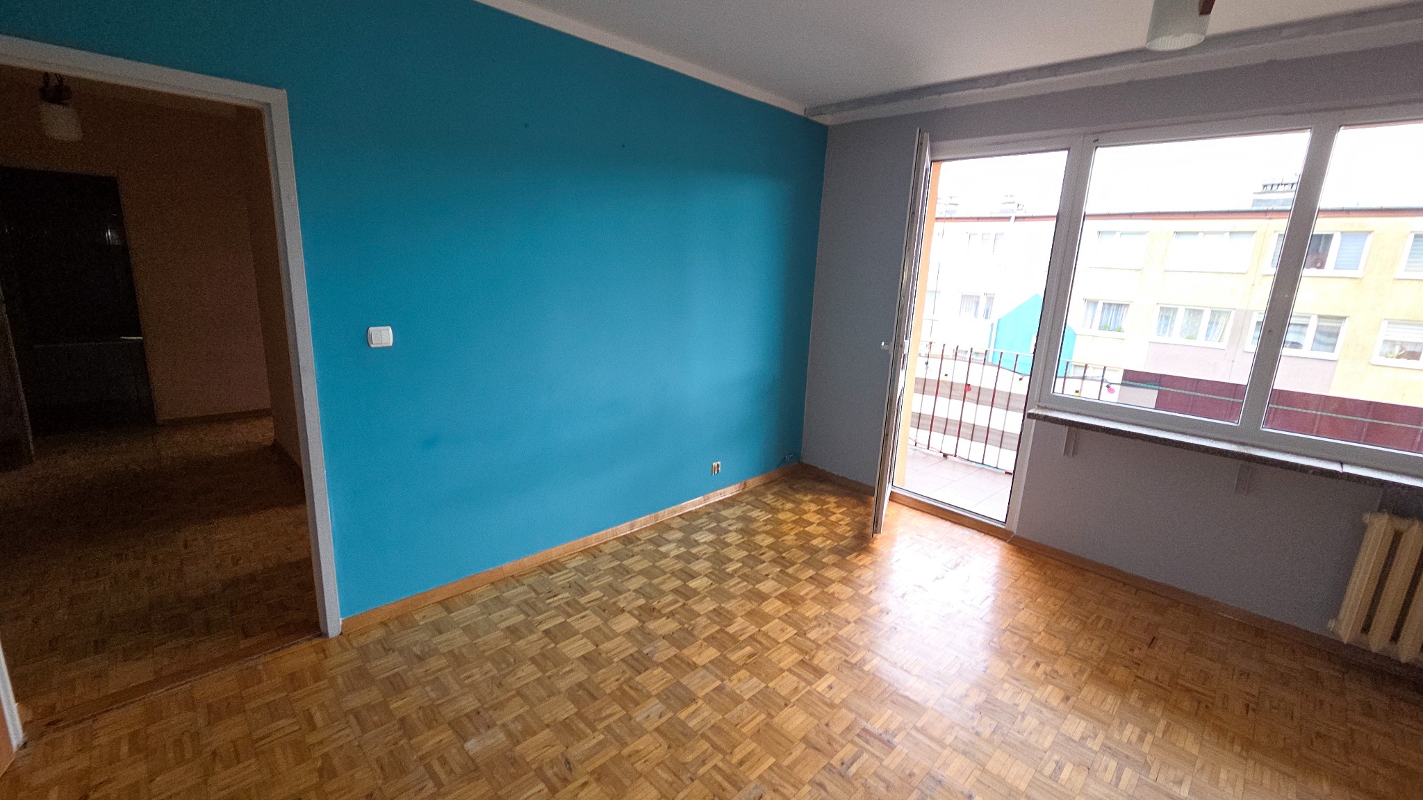 Pomalowane na turkusowo wnętrze pokoju z parkietem, widok na balkon i okno z widokiem na budynek. Świeżo odmalowane ściany, gotowe do dalszego wykończenia.