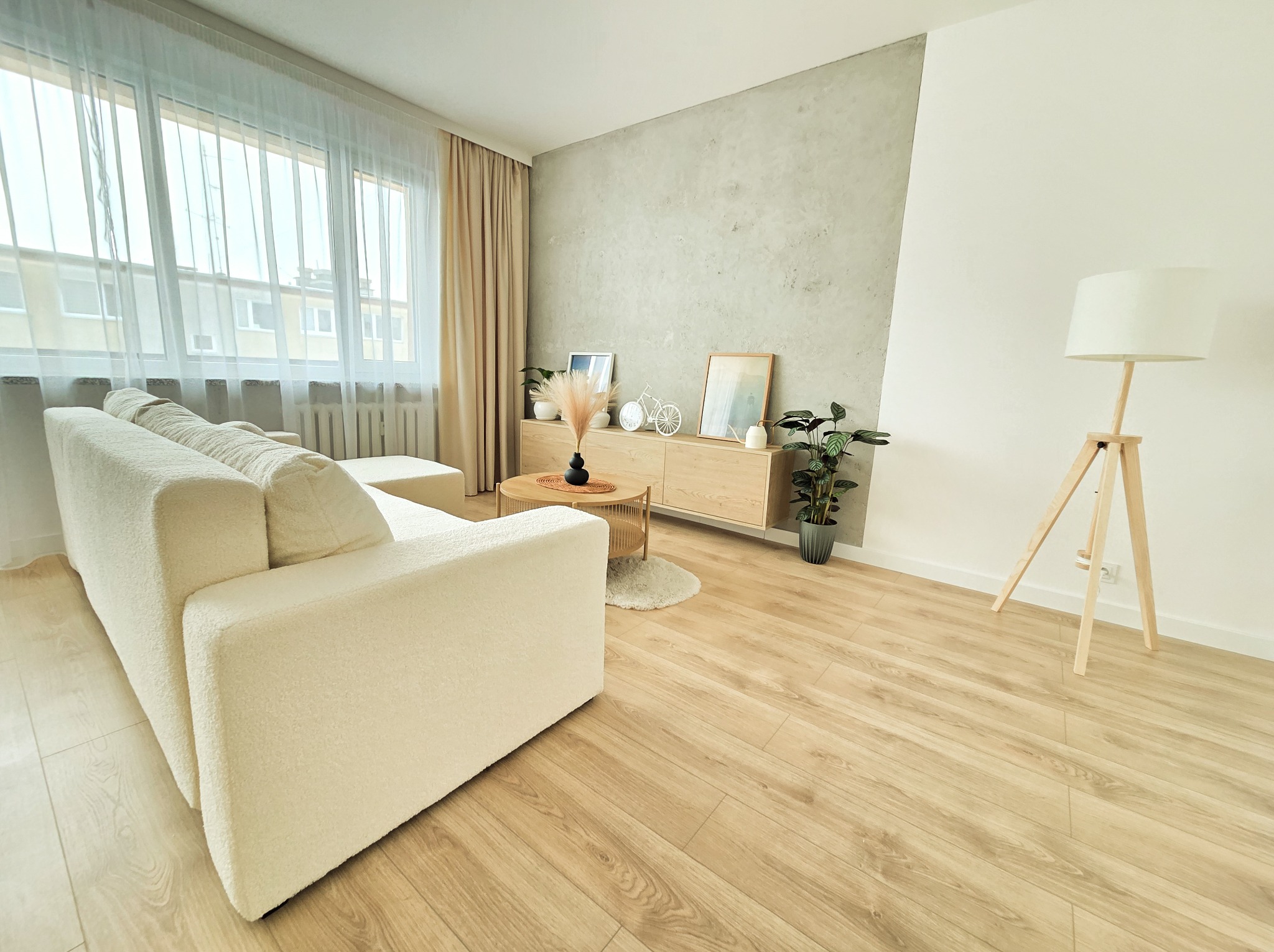 Jasny salon z drewnianą podłogą, kremową sofą i ścianą z efektem betonu. Widoczne okno z firankami, komoda z dekoracjami i lampa na trójnogu.