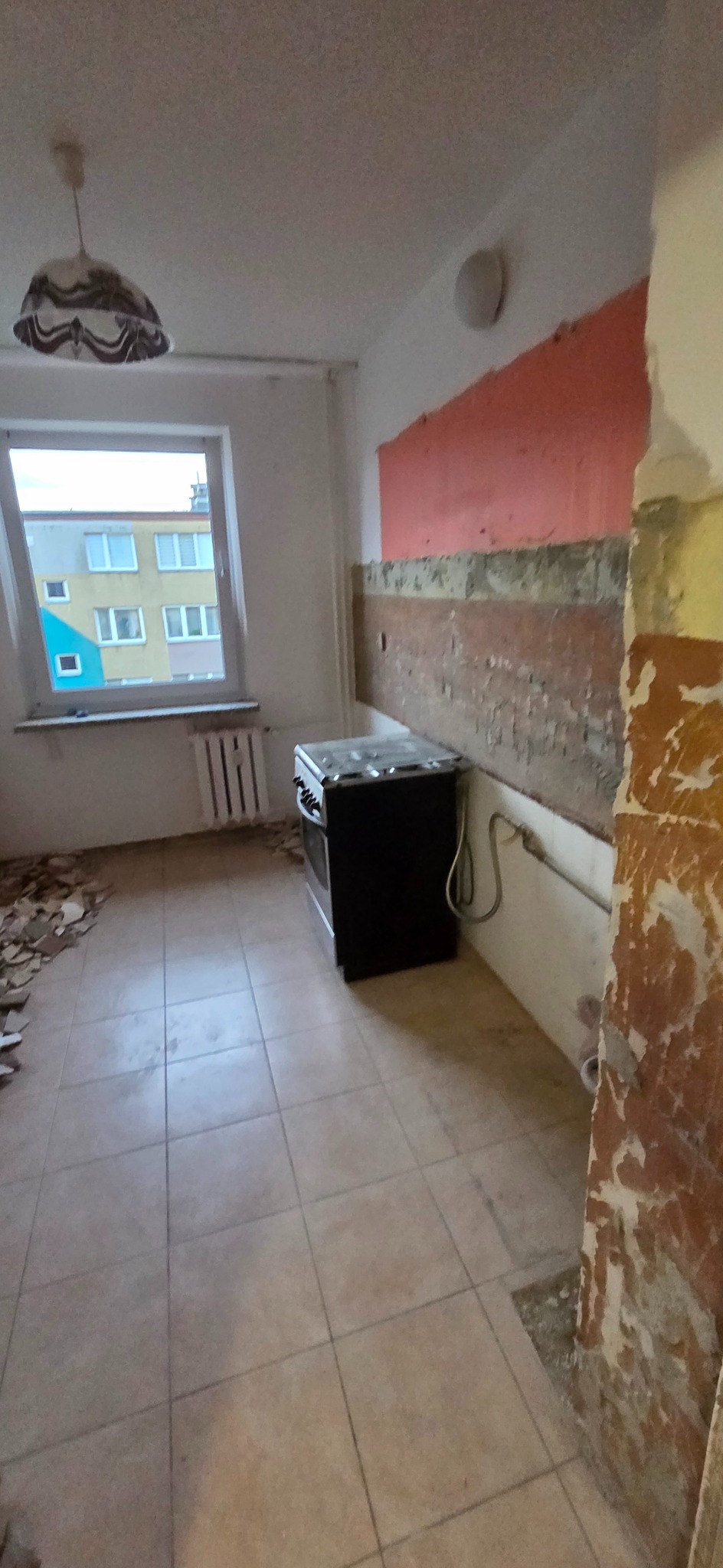 Kuchenne wnętrze w trakcie demontażu: odsłonięte warstwy farby na ścianach, częściowo zerwane płytki podłogowe, stara kuchenka. Widok z perspektywy wejścia.