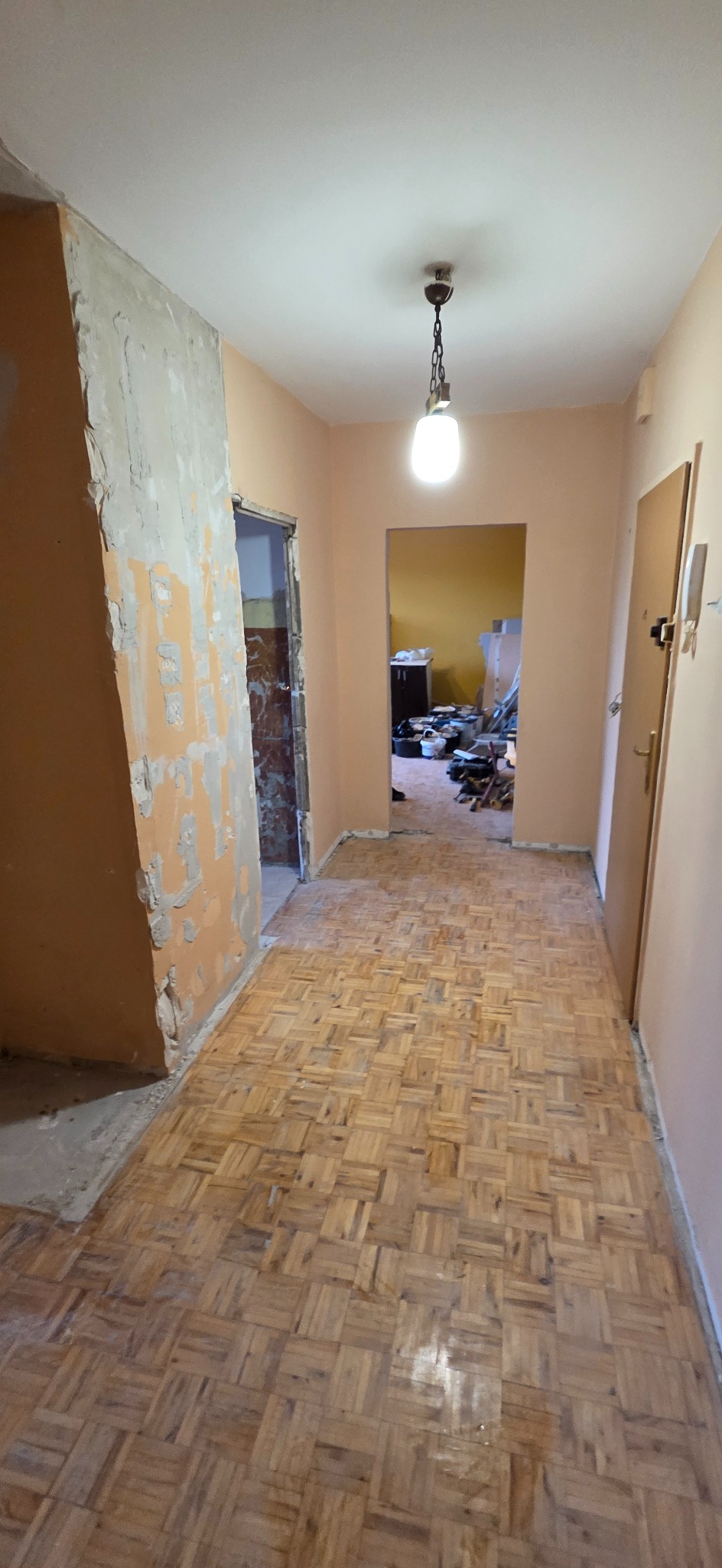 Korytarz w trakcie remontu: parkiet ułożony, ściany częściowo przygotowane do gładzi, widoczne ślady po starych powłokach. W tle widoczne pomieszczenie z narzędziami.